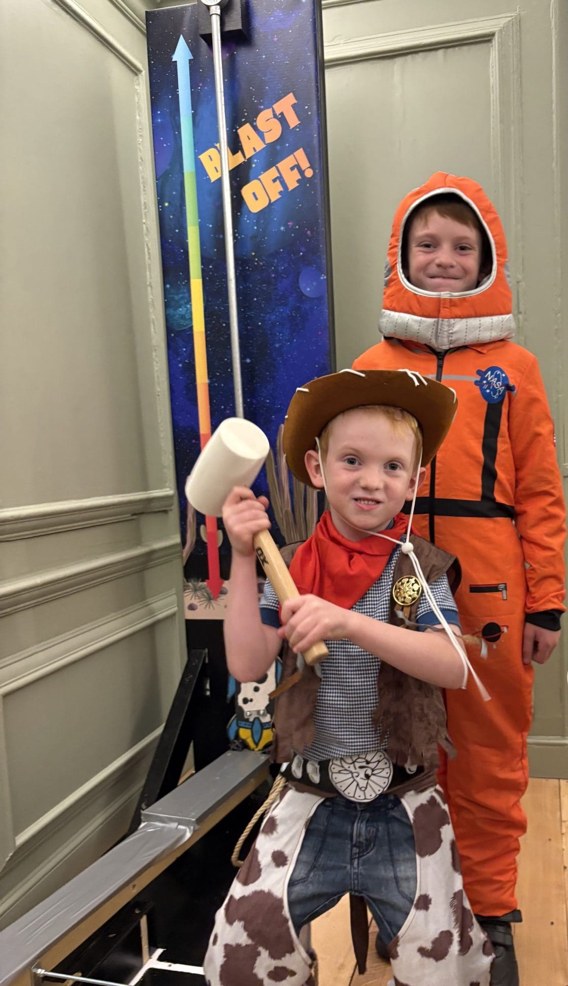 Two children stand in front of what looks like one of those old "Test your Strength" machines, where you hit a bar with a hammer as hard as you can and - if you're strong enough - a metal weight shoots up and dings a bell. One child is dressed as a cowboy and does indeed hold a wooden mallet. The other child is taller and is dressed in an orange spacesuit with a NASA badge on the chest.